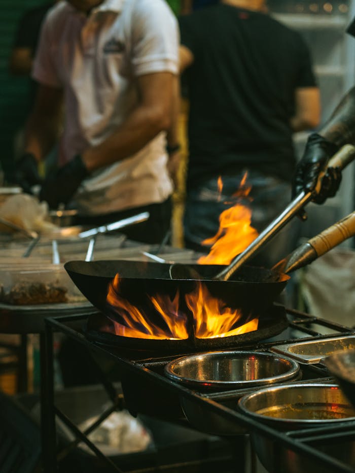 services-02 An energetic scene of Asian street food being cooked in a wok with flames on a bustling street.