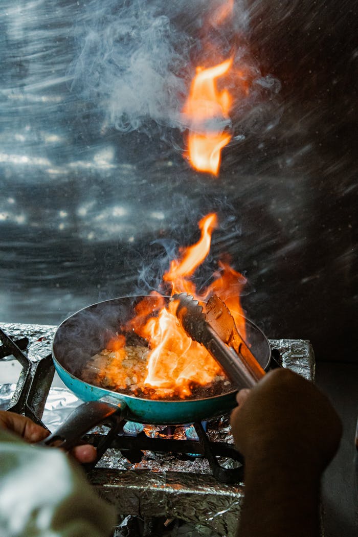 services-01 Chef using tongs to flambé dish on stove, dramatic fire for culinary skills.