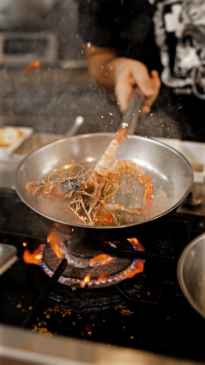 services-04 A chef expertly cooks prawns in a frying pan over a gas flame in a restaurant kitchen.